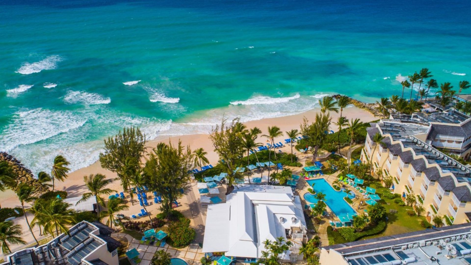 Aerial view of a tropical beach resort with turquoise ocean, palm trees, pools, and white buildings.