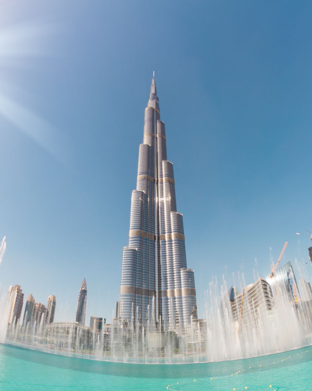 Burj Khalifa rises over turquoise fountains under a clear blue sky.