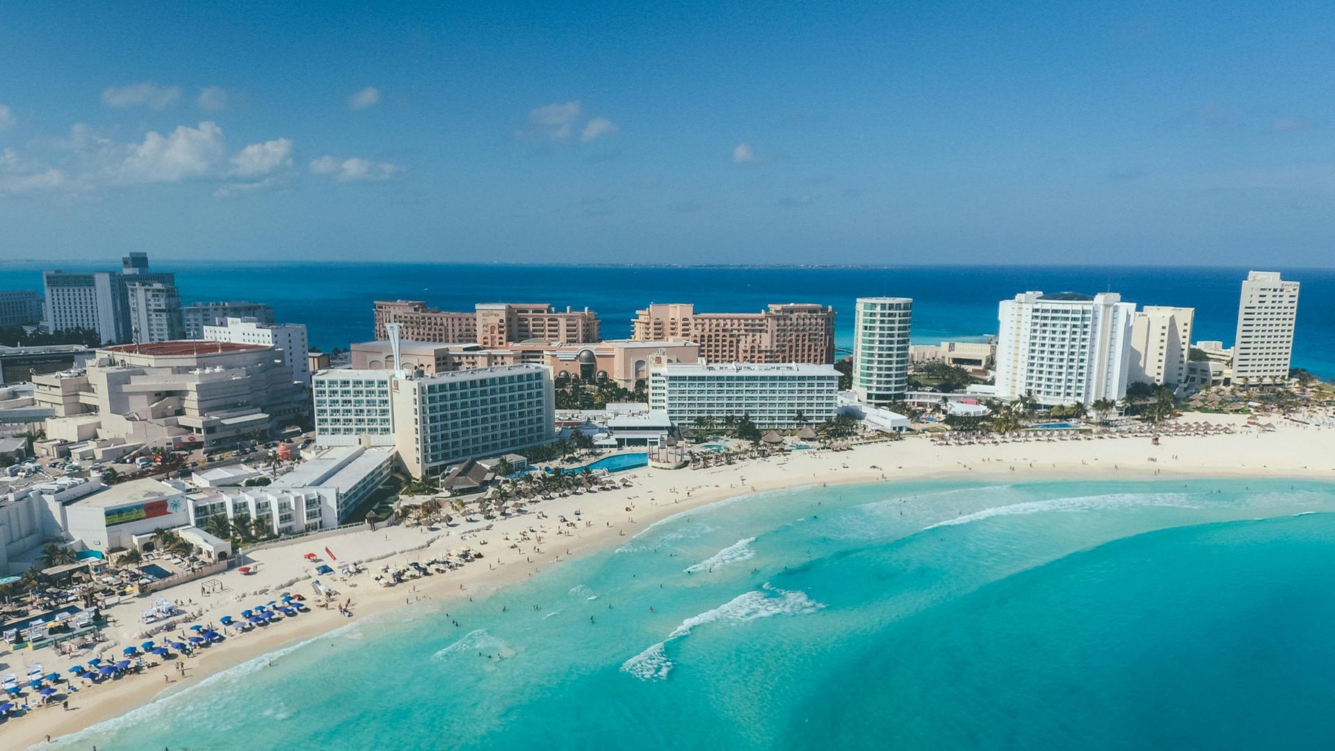 Aerial view of a turquoise beach with resorts, white sand, and calm ocean under a clear blue sky