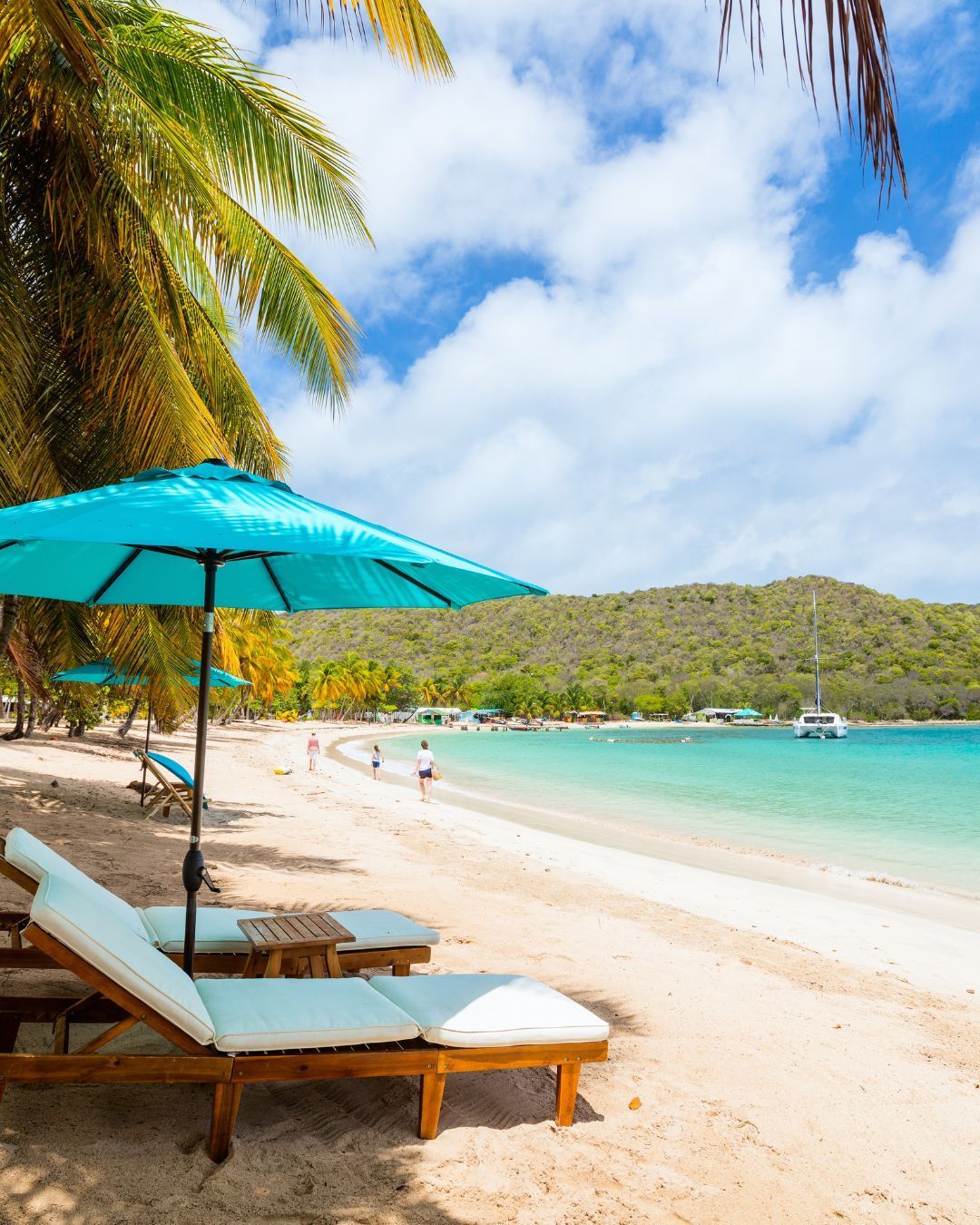 Tropical beach with turquoise water, white sand, palm trees, lounge chairs, and a sailboat offshore