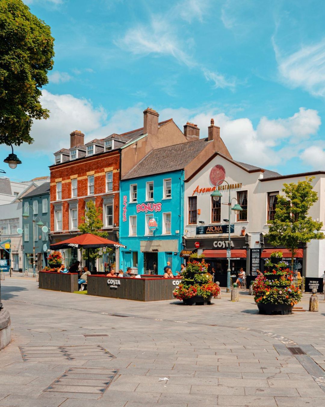 Colorful storefronts and flower planters lining a quiet European-style town square under a bright blue sky