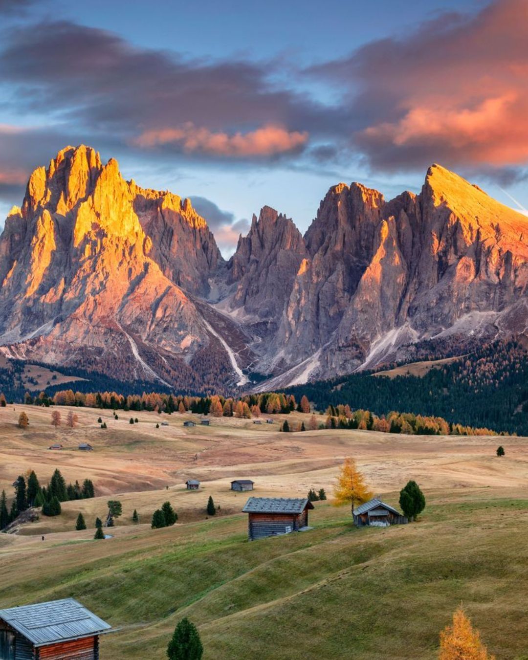 Sunlit jagged mountains above rolling golden meadows with a few small cabins at sunset