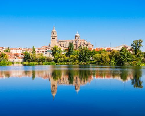 City skyline with cathedral towers reflected in a calm blue river under a clear sky