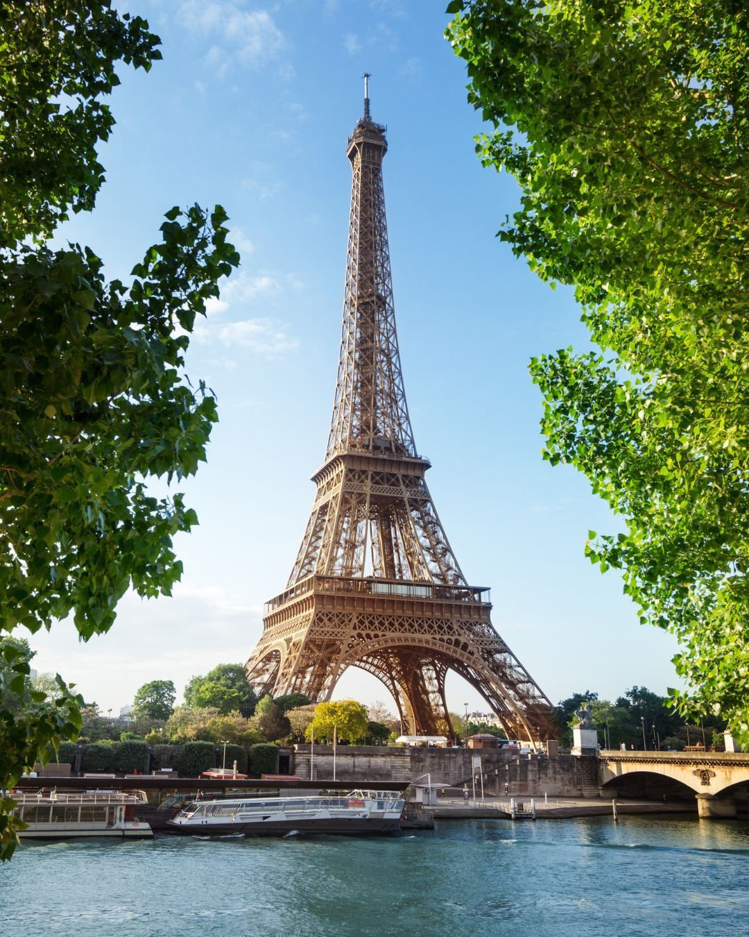 Eiffel Tower framed by green trees beside the Seine under a clear blue sky