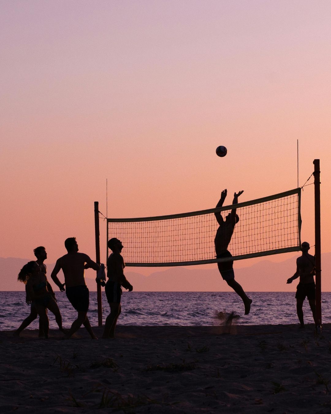 Silhouetted beach volleyball players at sunset, one leaping to hit the ball over the net.