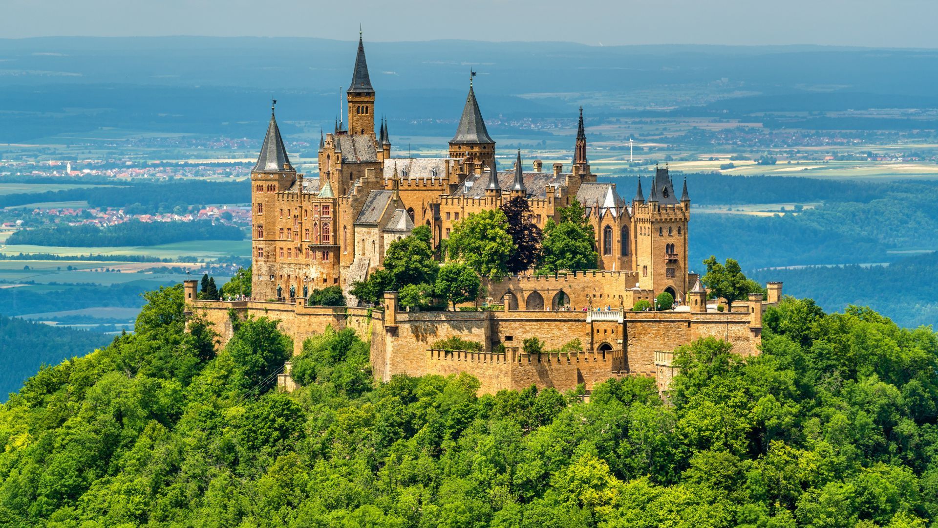 Castle Hohenzollern atop a green, tree-covered hill with a scenic mountain backdrop.