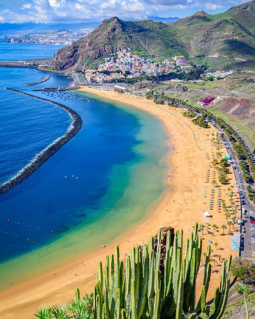 Aerial view of a tropical bay with golden beach, blue water, mountains, and a coastal town.