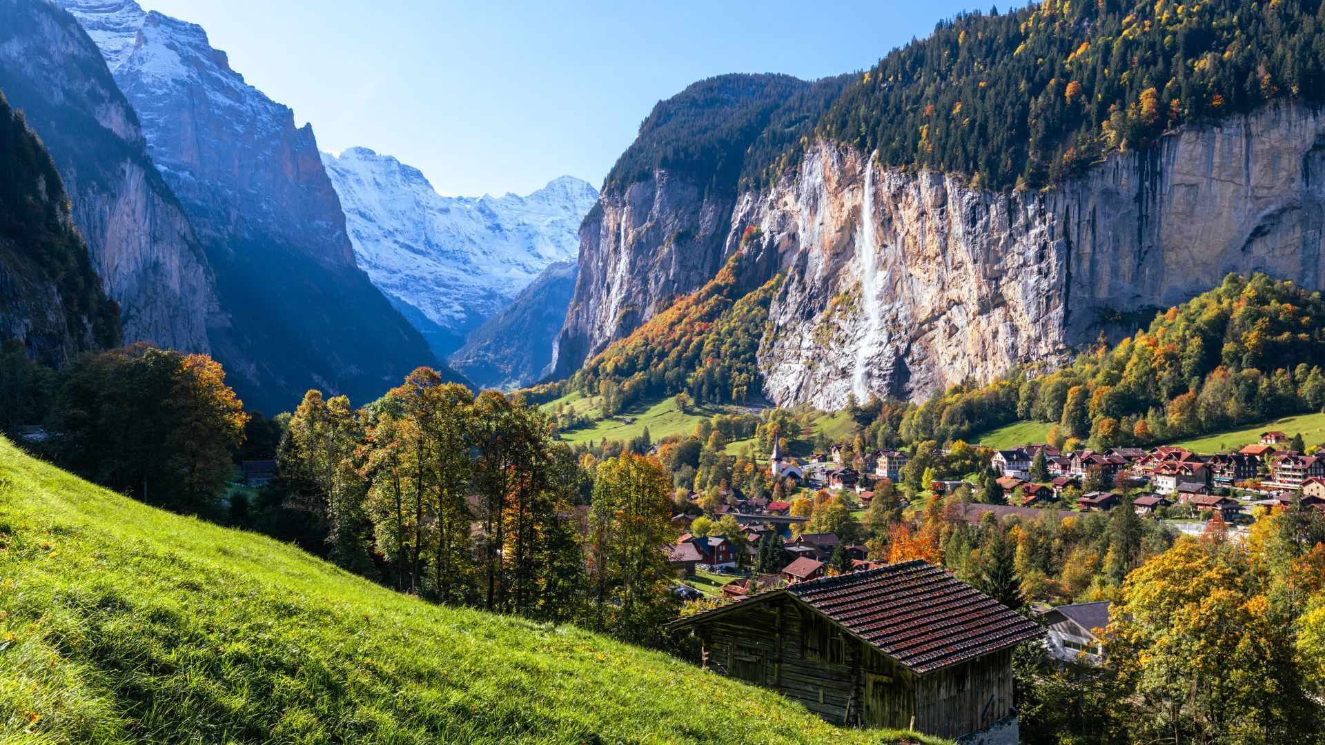 Green valley with village, waterfalls, snowy mountains, and autumn trees under blue sky.