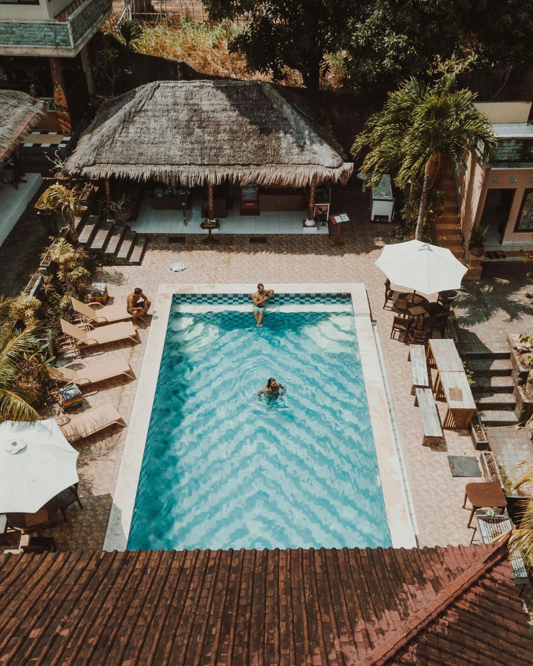 Aerial view of a resort pool with one swimmer, lounge chairs, and thatched-roof buildings