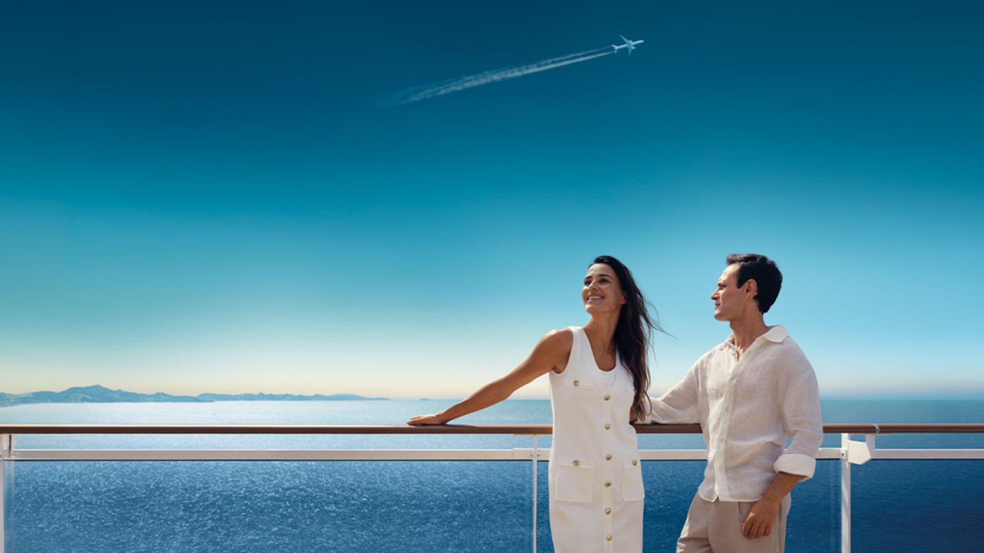 Two people on a balcony overlooking the ocean under a bright blue sky, smiling and holding hands.