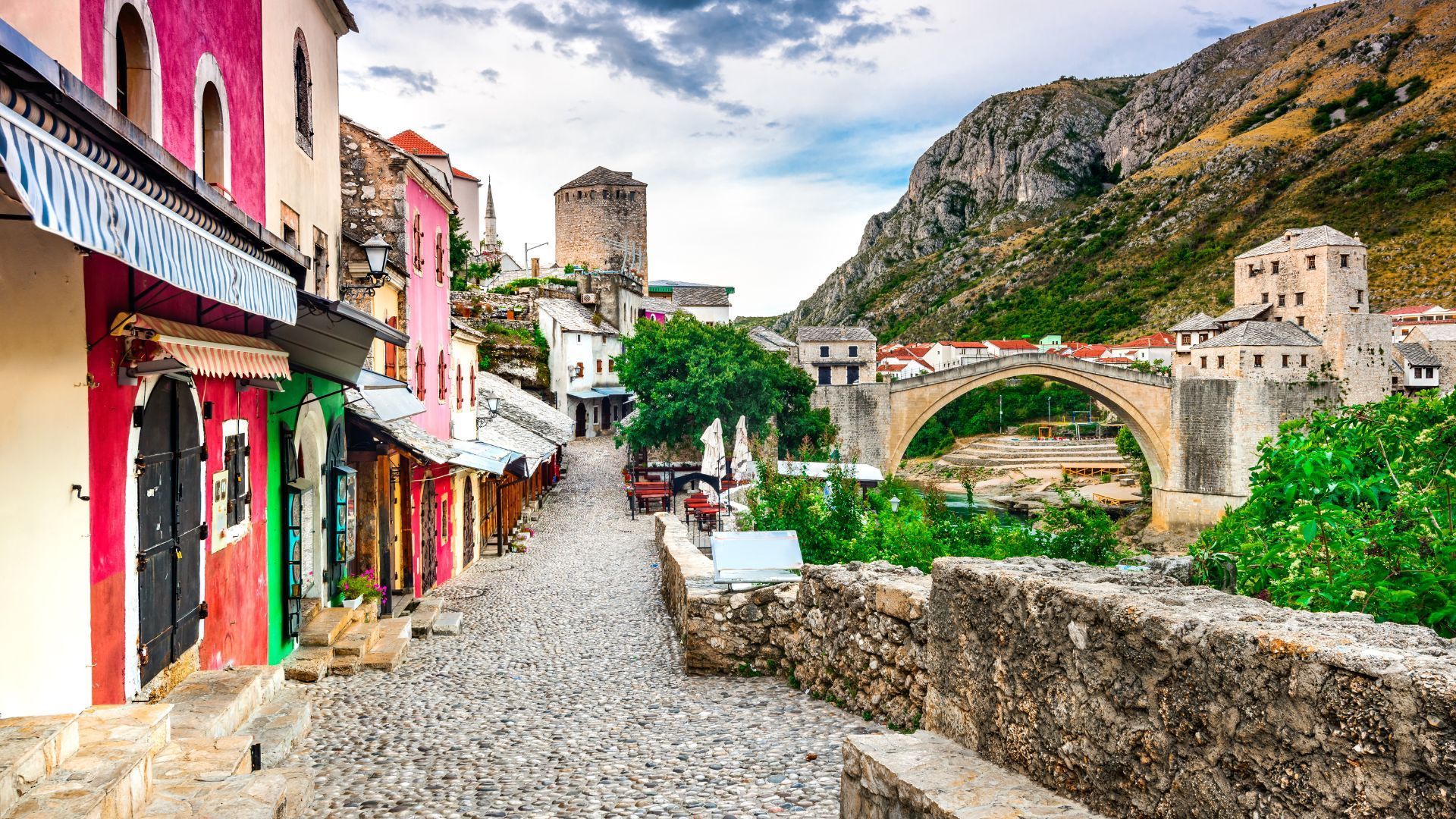 Cobblestone street lined with colorful shops leading to a historic bridge and mountains in Mostar, Bosnia and Herzegovina.