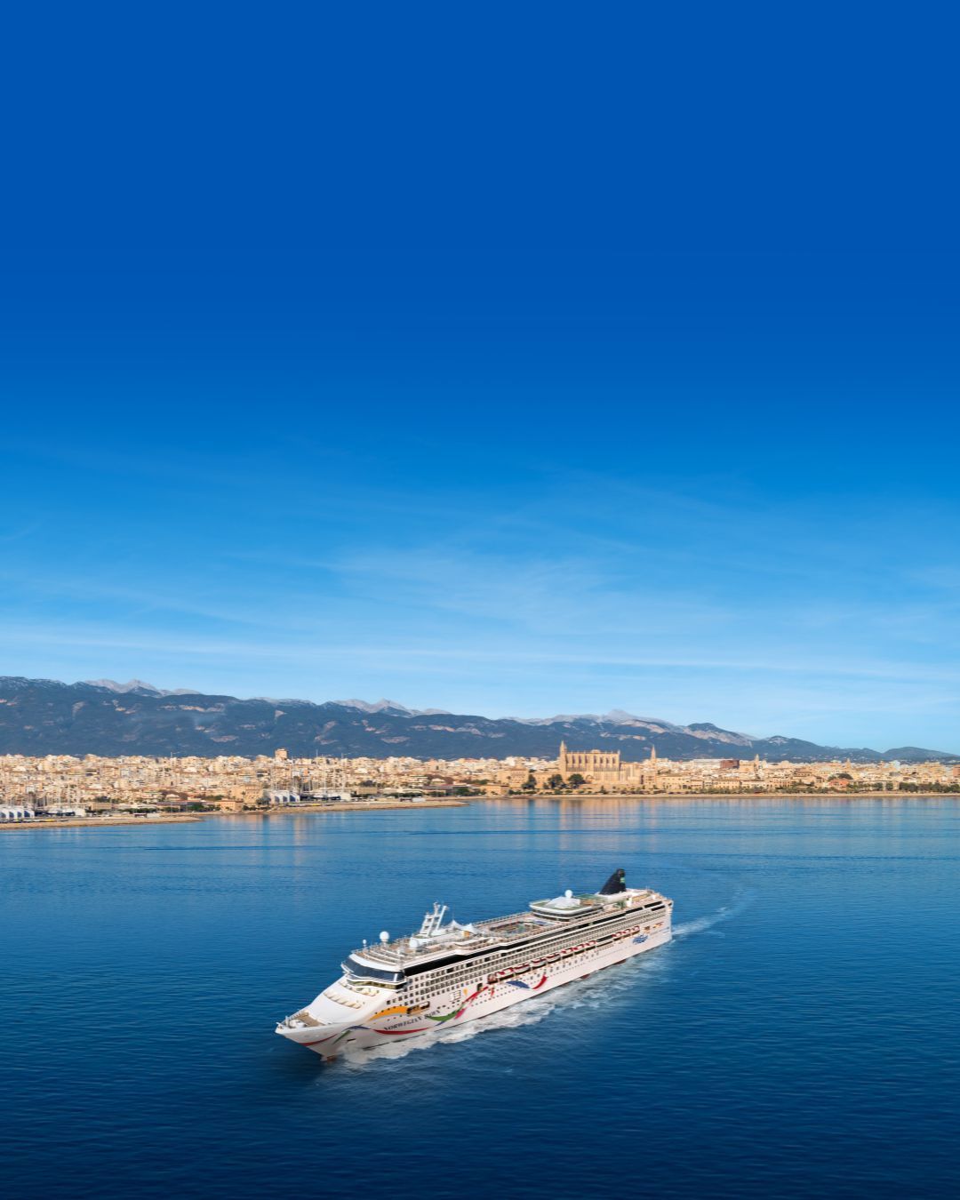 White cruise ship from Norwegian Cruise Line on calm blue water with a coastal city and mountains under a clear sky
