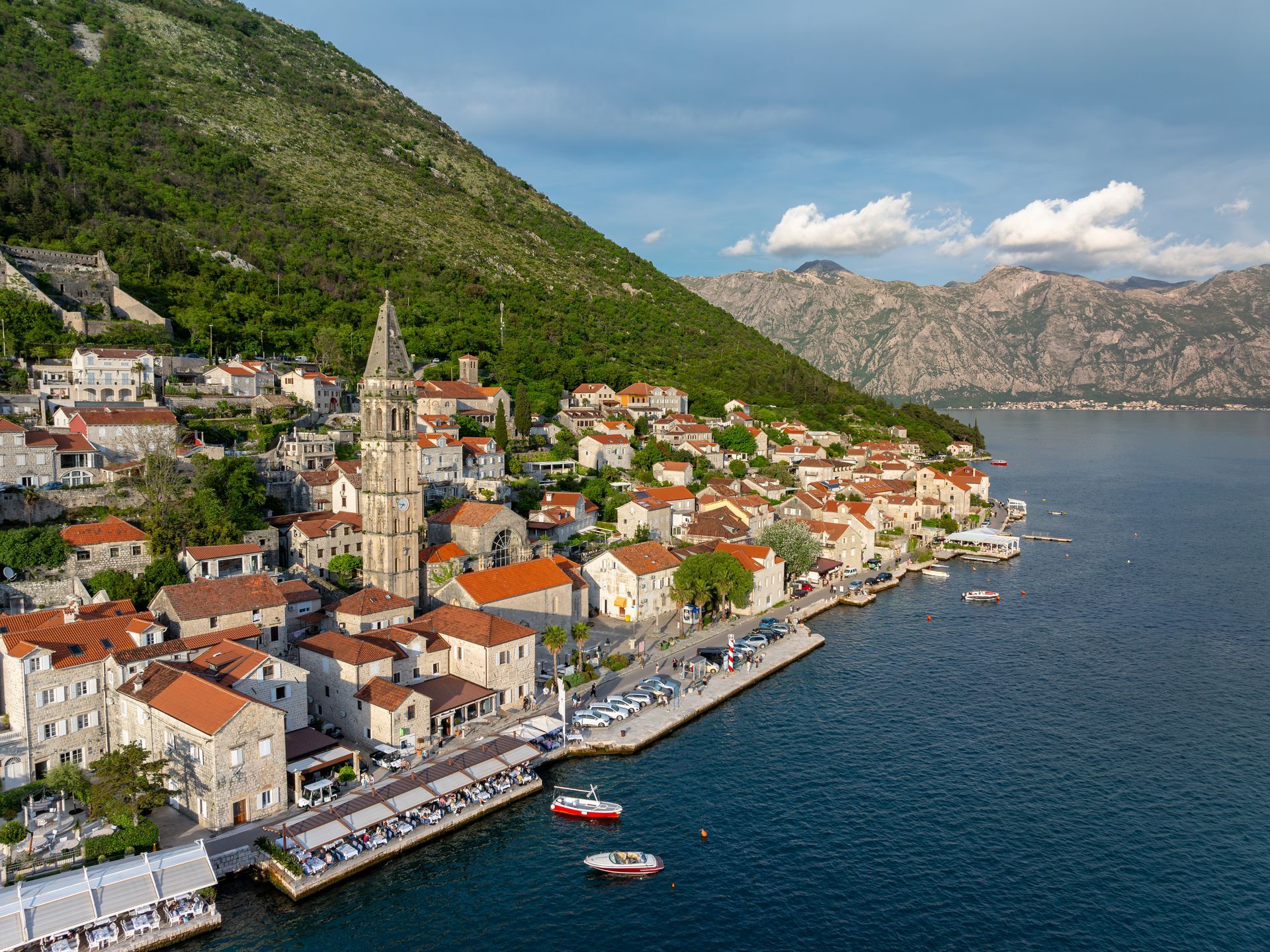 Coastal town Kotor in Montenegro with red-roofed buildings, church tower, and harbor beside a steep green hillside and blue sea