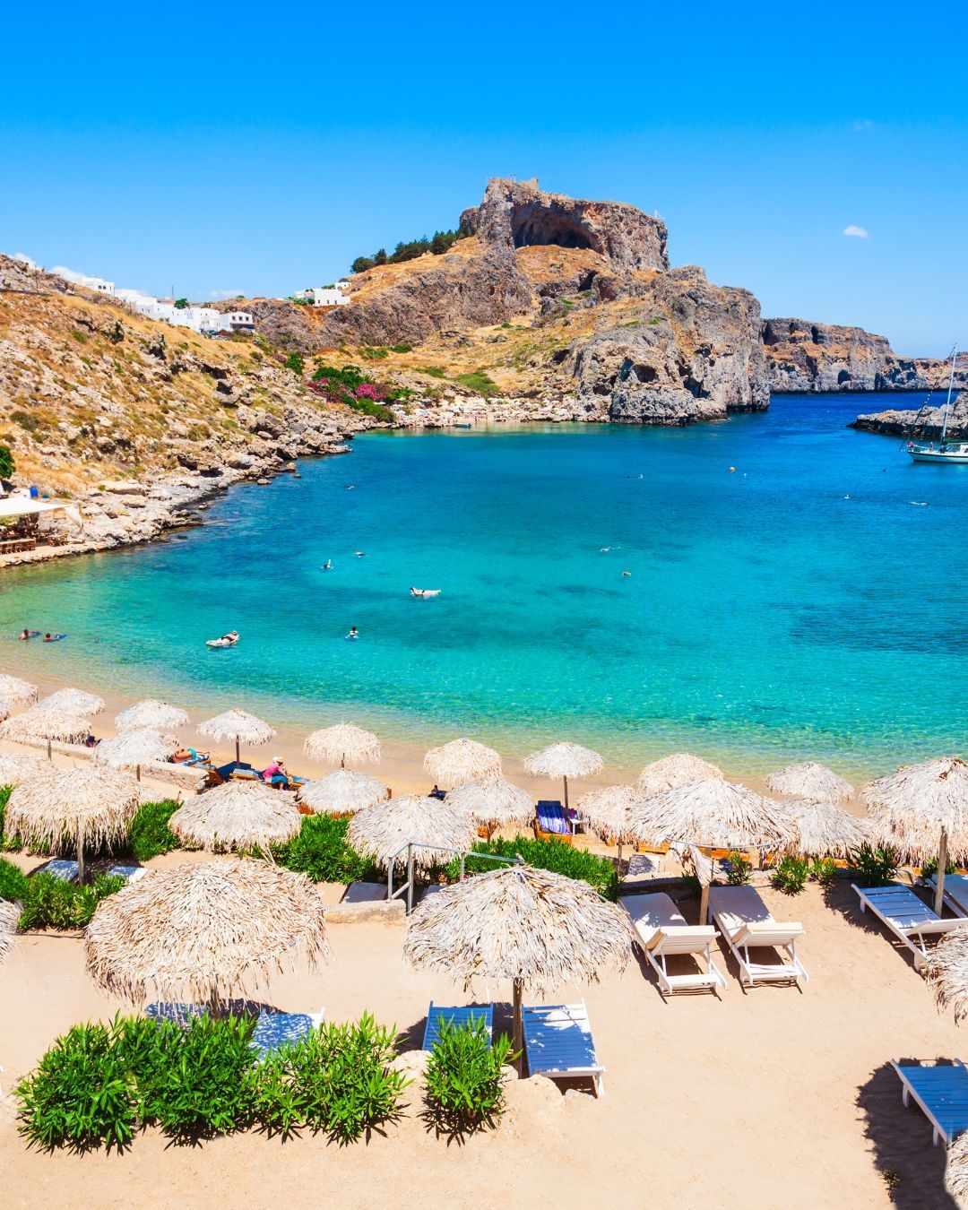 Turquoise cove with sandy beach, thatched umbrellas, lounge chairs, and rocky cliffs under a blue sky