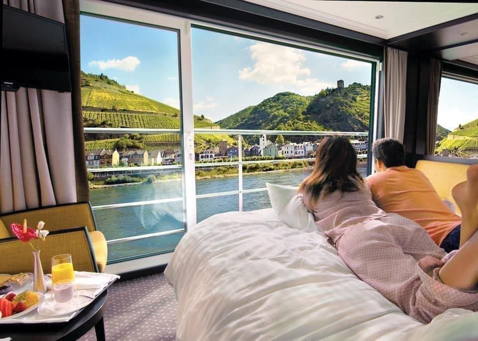 Couple on a bed overlooking a bright pool and green hills through a large window, Avalon Waterways