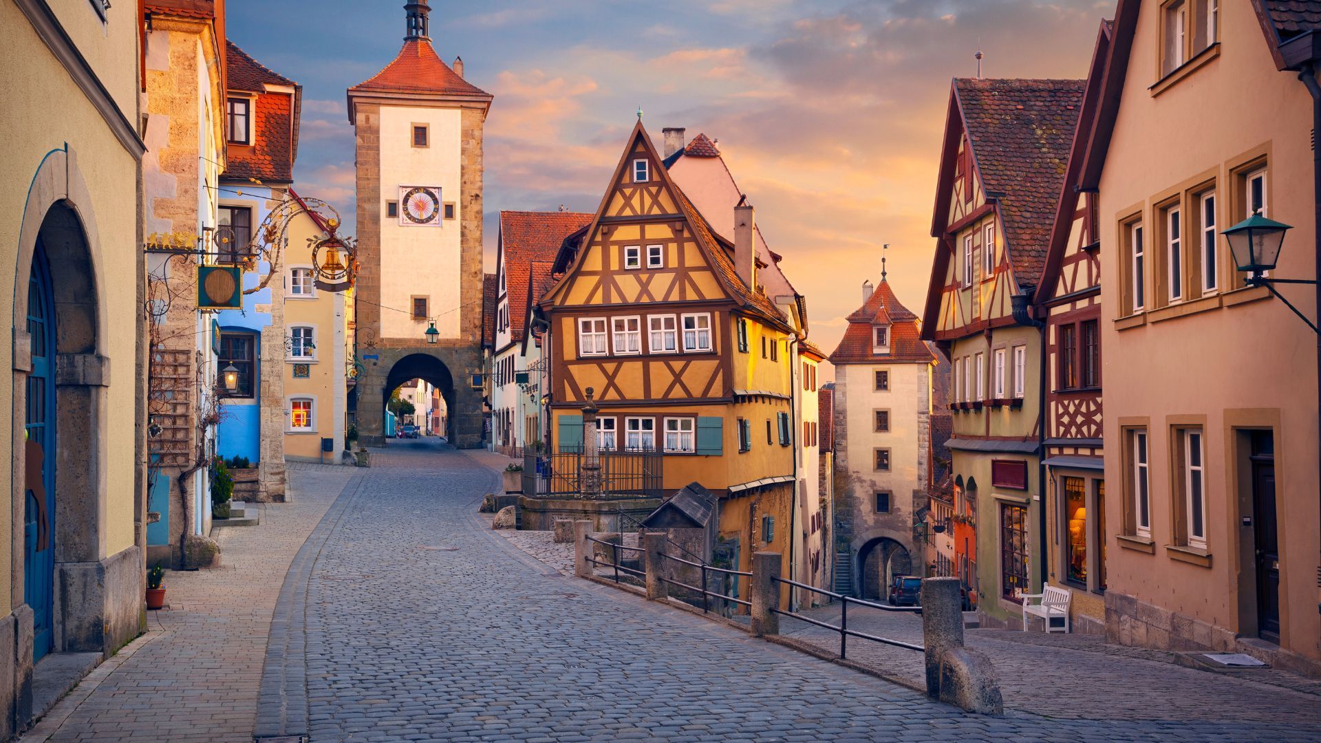 Cobblestone street lined with colorful, historic buildings, towers, and archway in a European town at sunset.