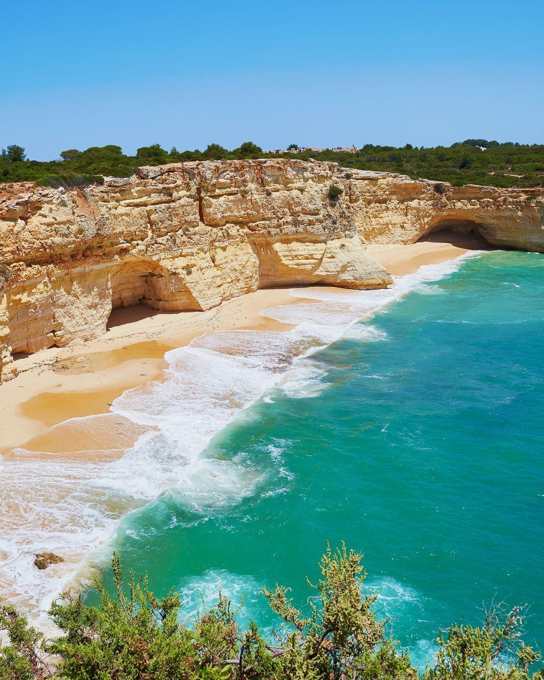 Turquoise waves lapping a sandy beach below sunlit rocky cliffs under a clear blue sky