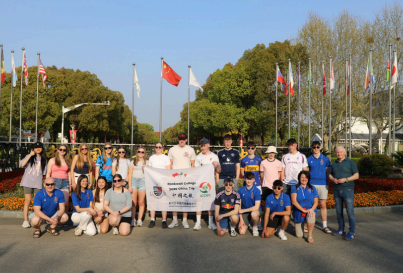 Large student's group of students posing outdoors in front of flags, holding a banner on a sunny day.