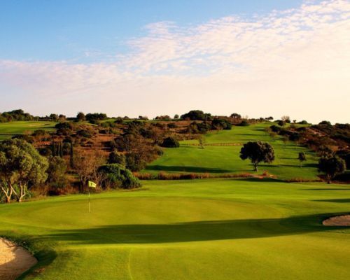 Sunlit golf course with rolling green fairways, scattered trees, and a partly cloudy sky.