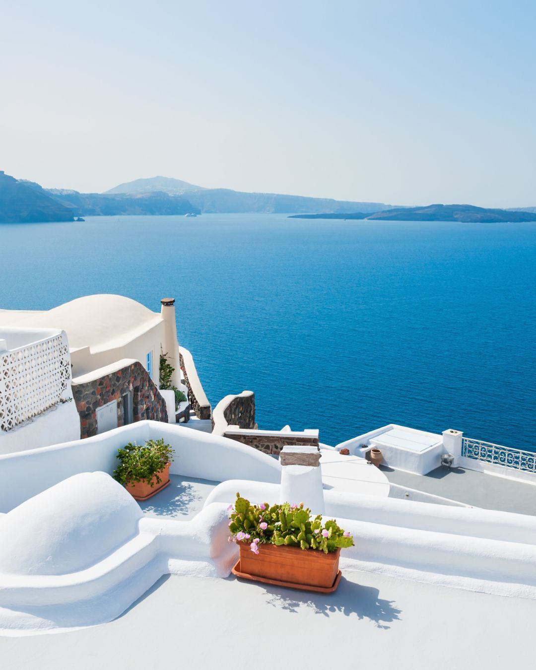 White terrace overlooking a bright blue sea with distant islands and potted flowers in the foreground