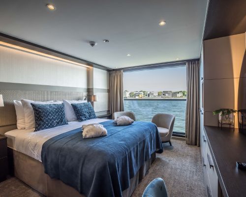 Travelmarvel Vega river cruise ship, bedroom with blue bedspread, two towels, and a large window overlooking water