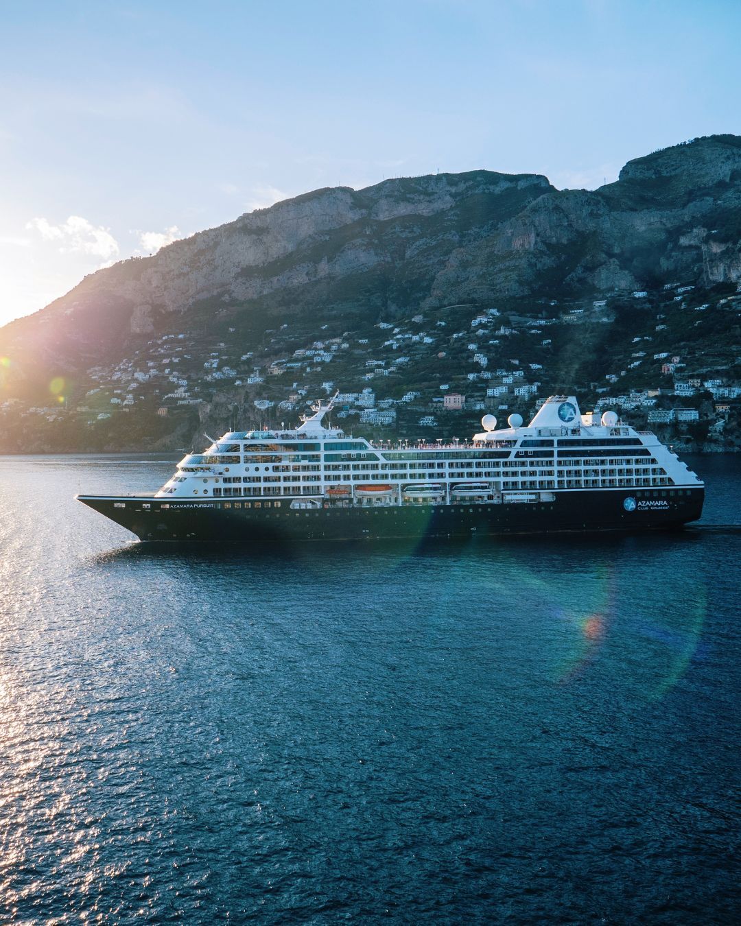 Cruise ship Azamara Pursuit on calm blue water beside a rocky coastal hillside at sunset