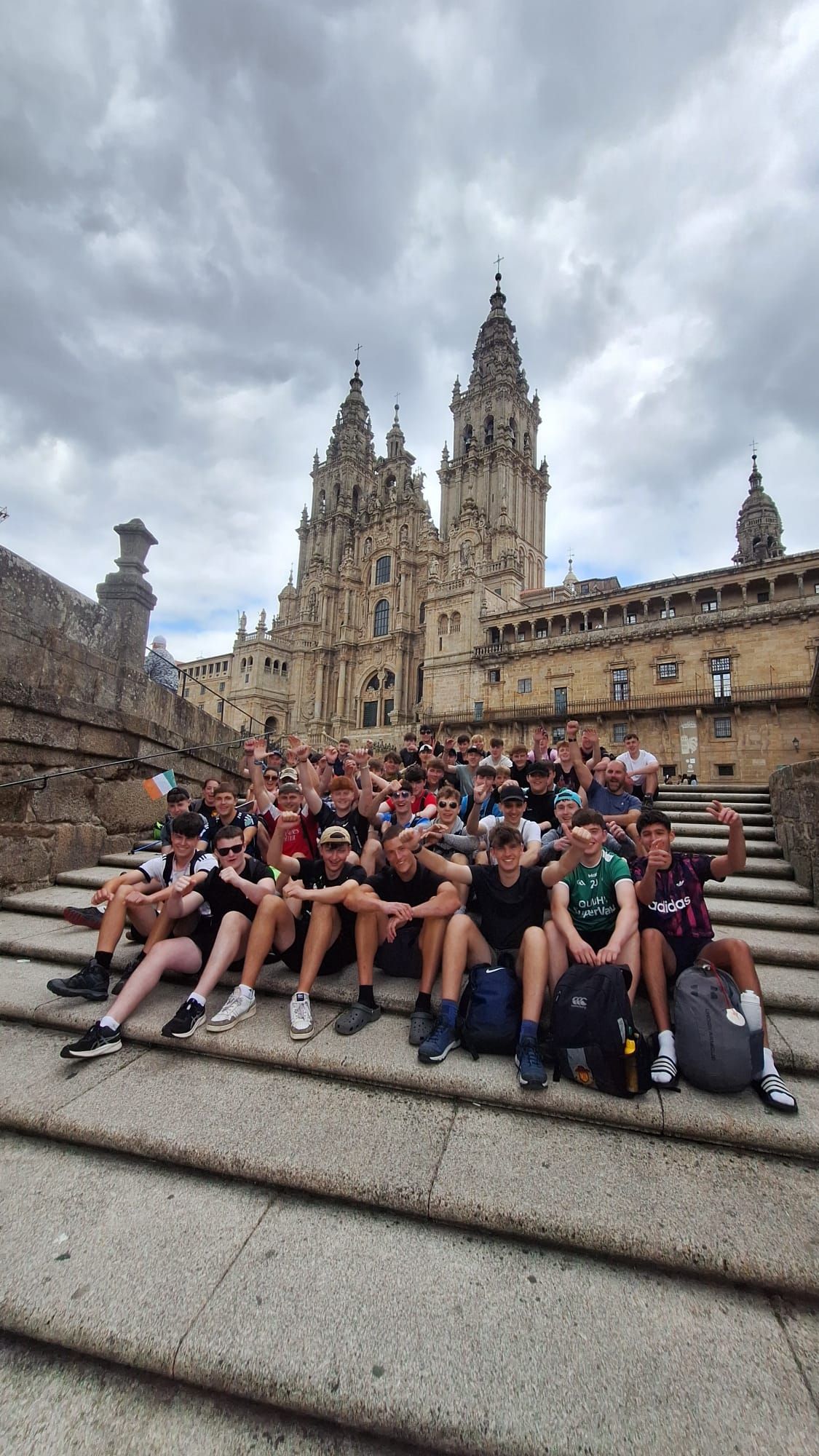 Group of tourists sitting on cathedral steps under a cloudy sky, with the ornate façade behind them.