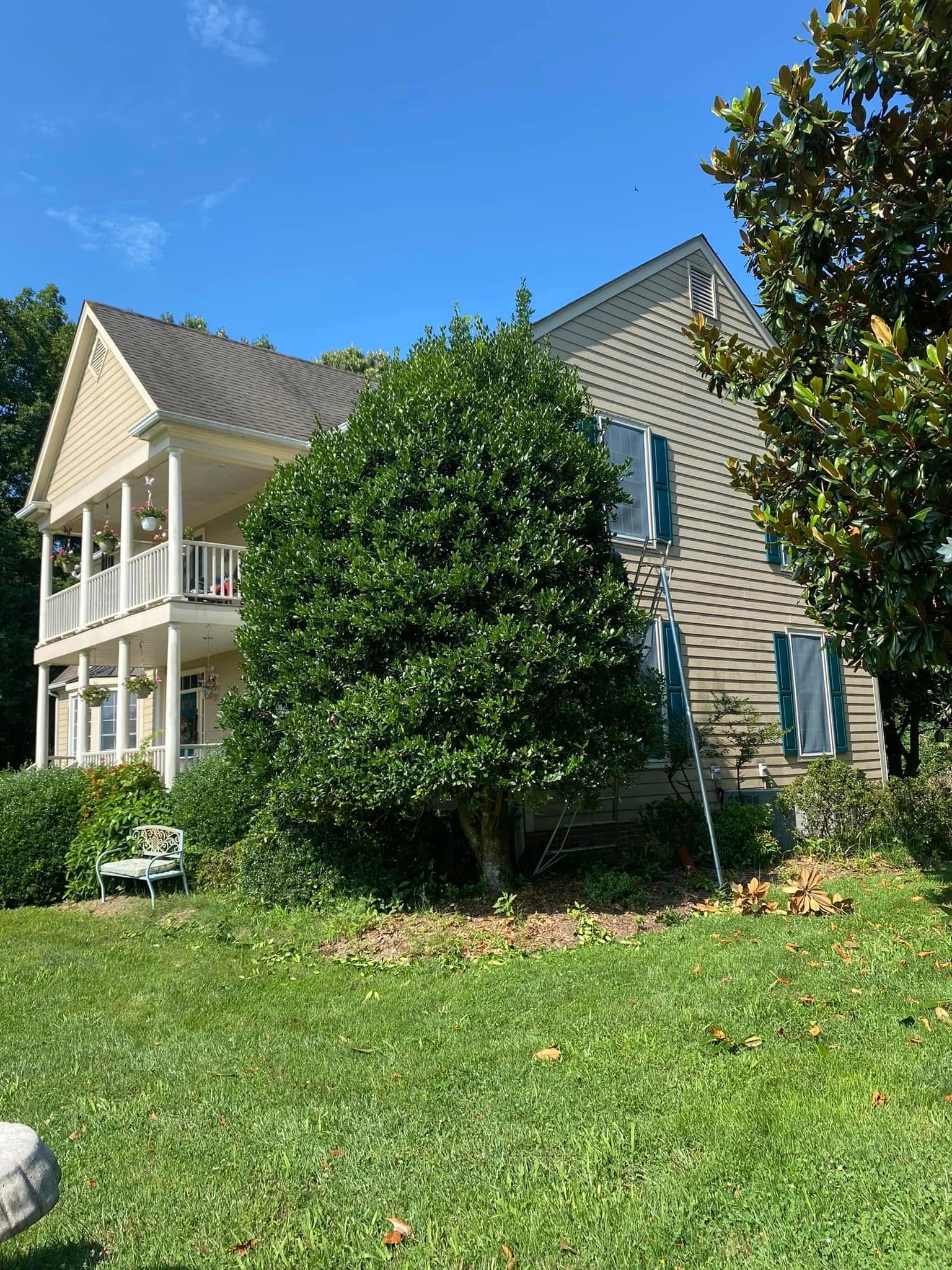 A two-story light-colored house with a wraparound porch, surrounded by lush green trees and a lawn under a blue sky.