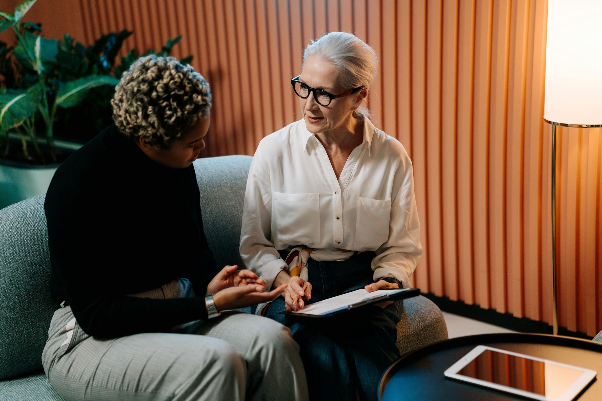 Two women in a conversation, one holding a tablet. Seated on a couch, in a room with pink walls.