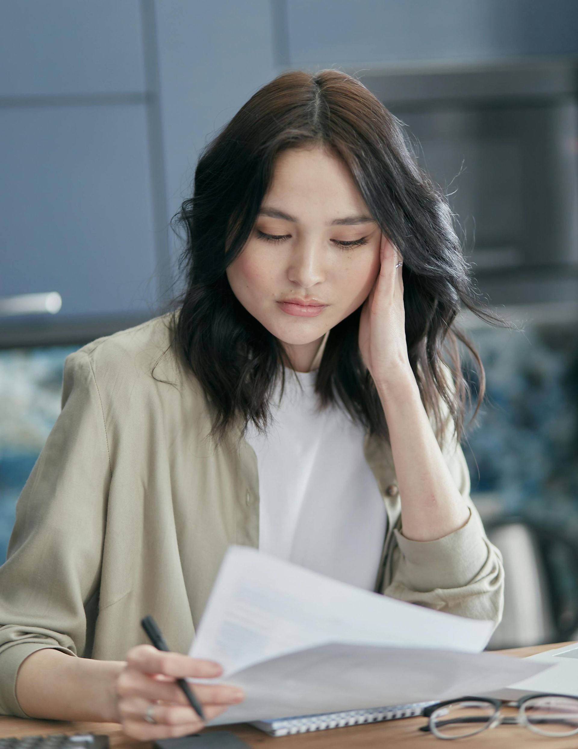 Woman, hand on head, looking at papers. Beige jacket, white shirt, kitchen background.