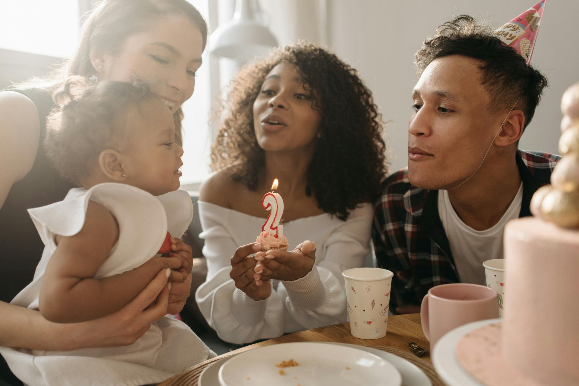 Family celebrating a toddler's birthday. Blowing out a candle, everyone smiles, indoors, near a cake.