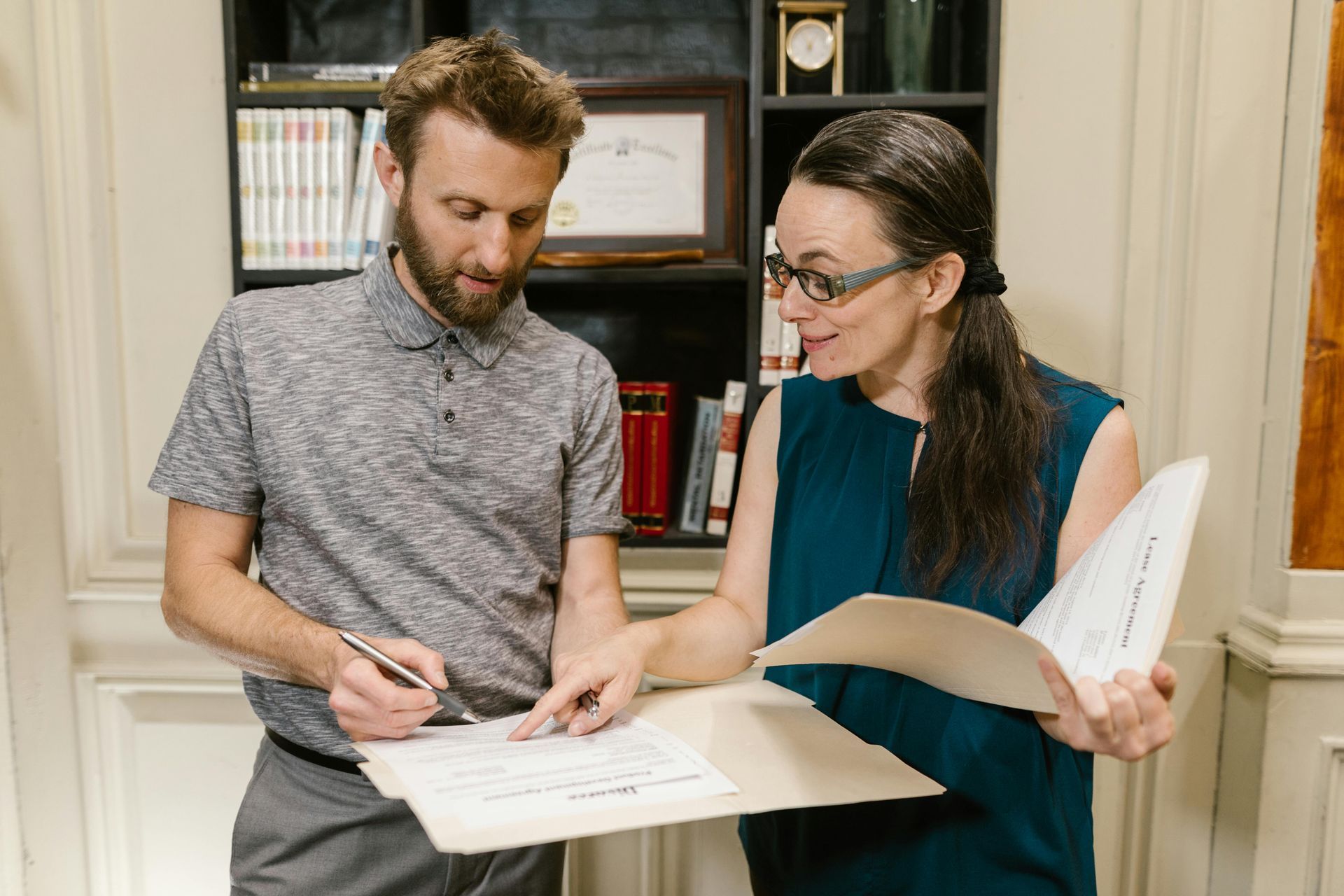 Man and woman reviewing papers in an office setting; man points with pen, woman wears glasses.
