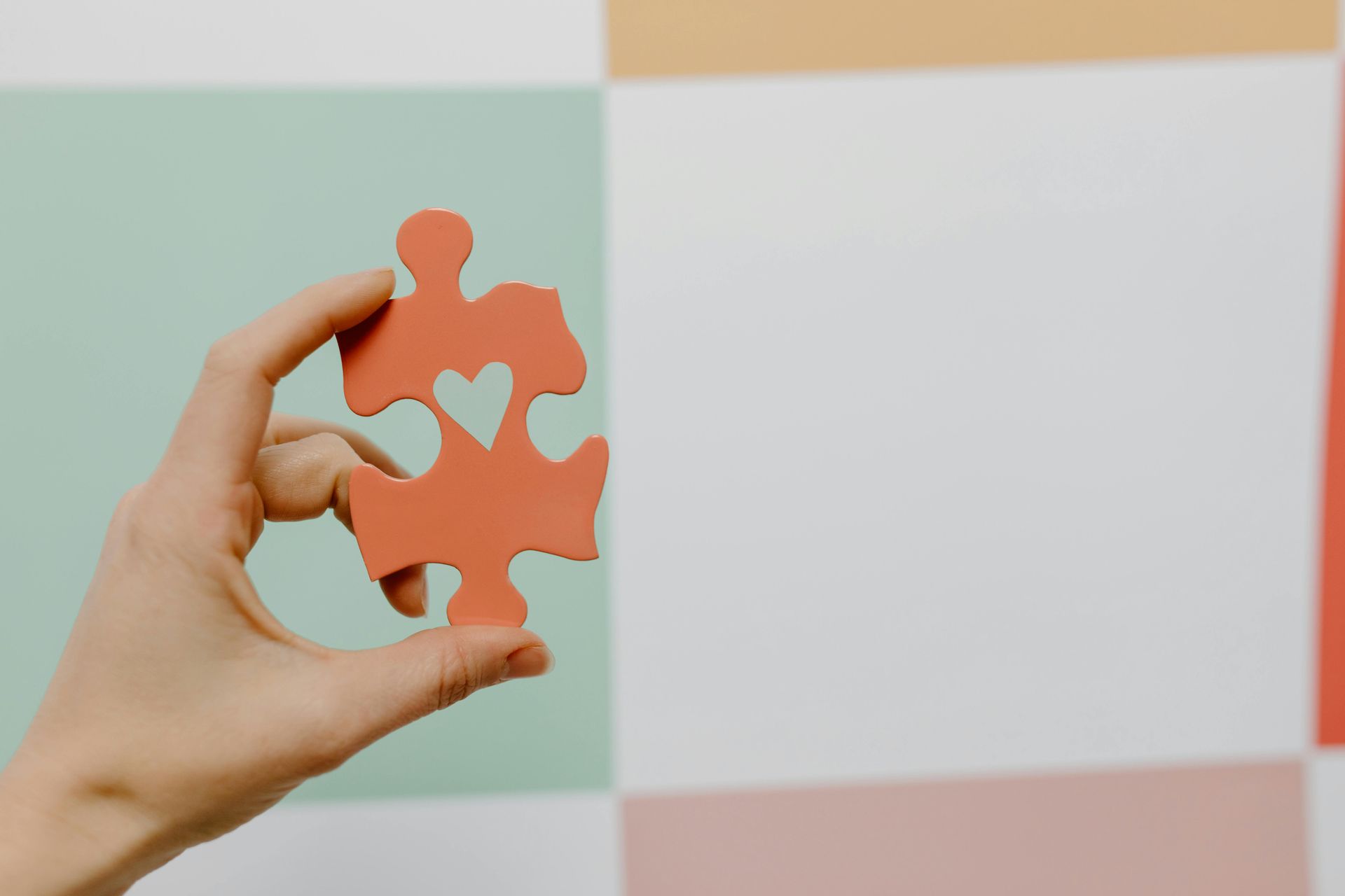 Hand holding a coral jigsaw puzzle piece with a heart-shaped cutout, against a pastel, checkered background.