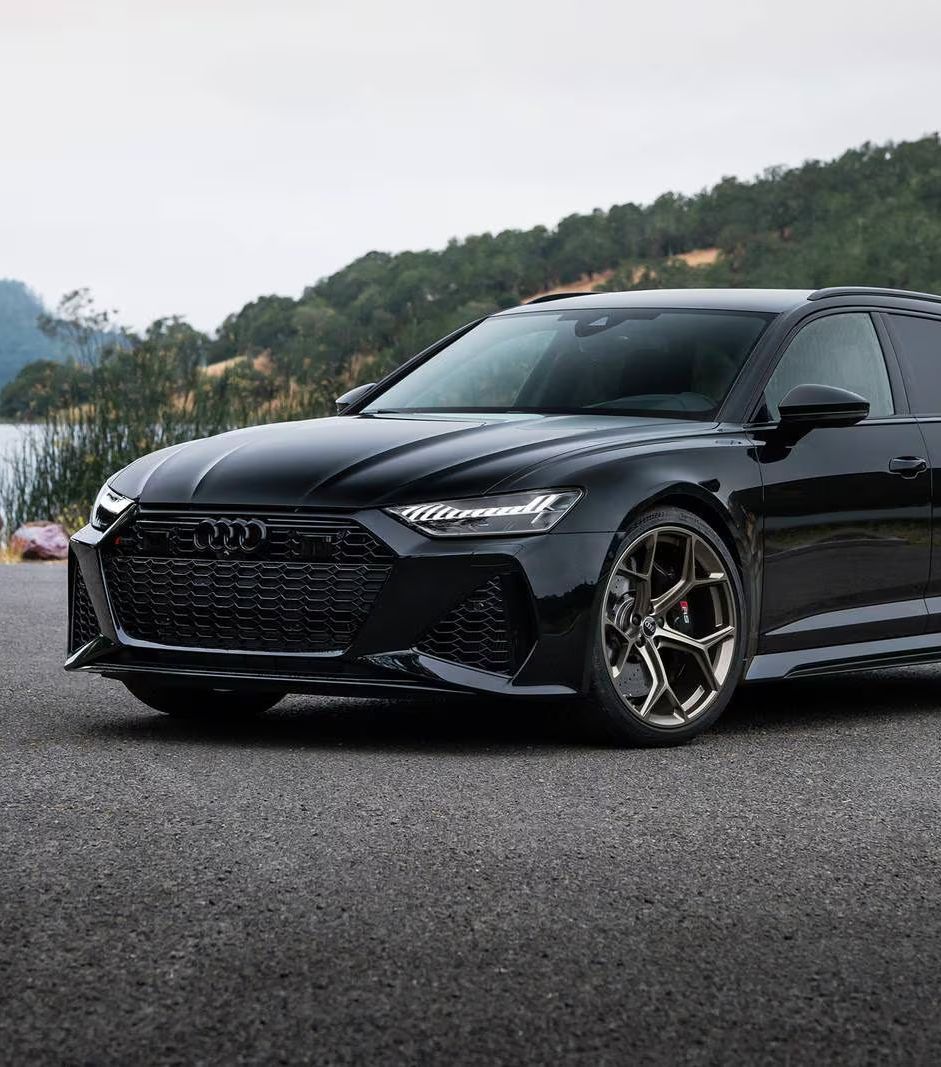 Black Audi RS6 Avant parked on asphalt road, with a lake and trees in the background.