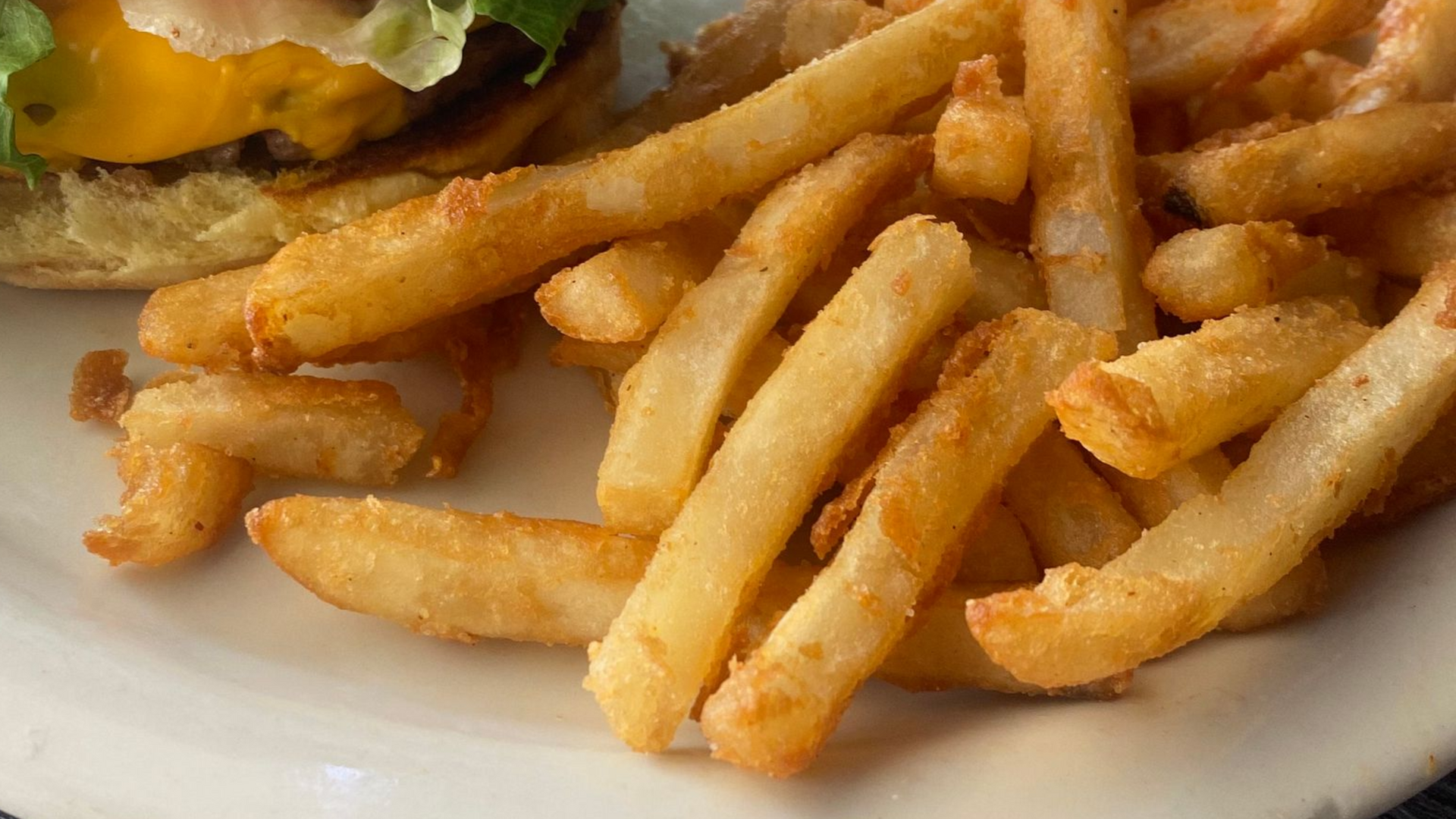 Plate of golden-brown french fries, with part of a burger visible in the background.