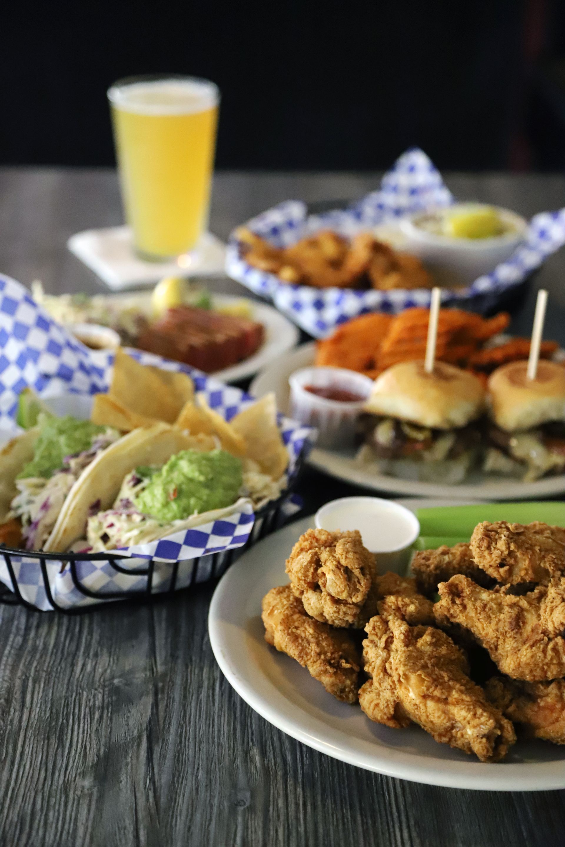 Assorted bar food: wings, sliders, tacos, onion rings, sweet potato fries, beer, on a table.