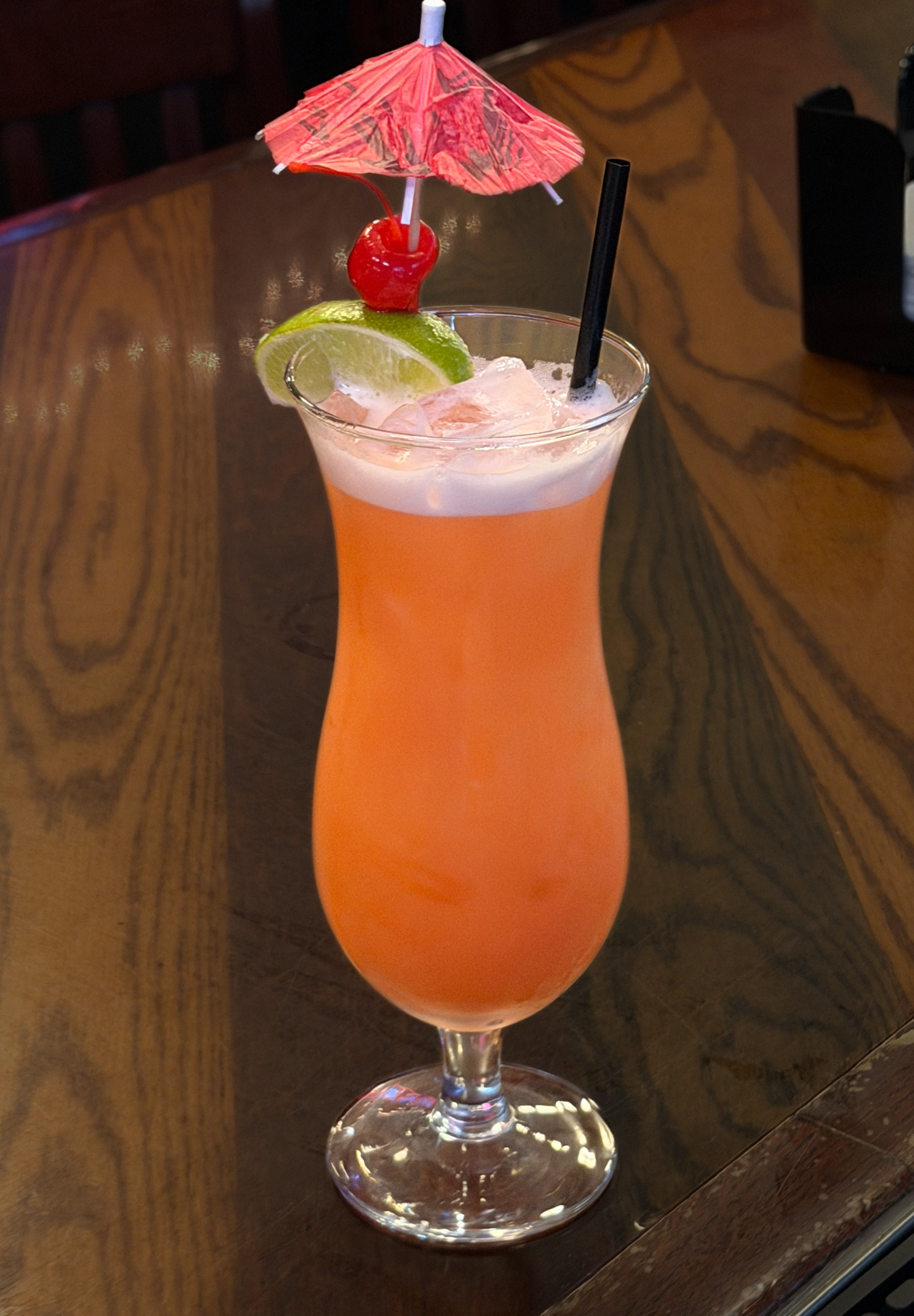Orange tropical cocktail in a tall glass with a lime slice, cherry, pink umbrella, and straw on a table