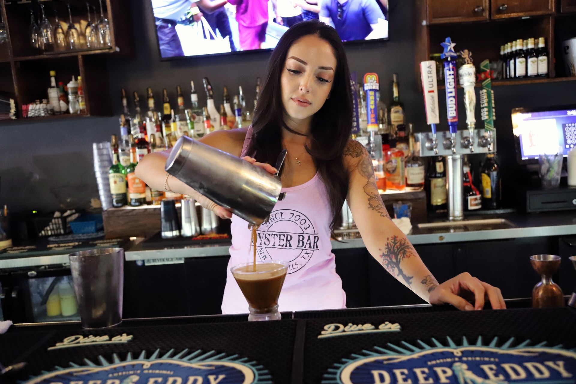 Bartender pouring a dark drink into a martini glass, behind a bar with bottles and taps.