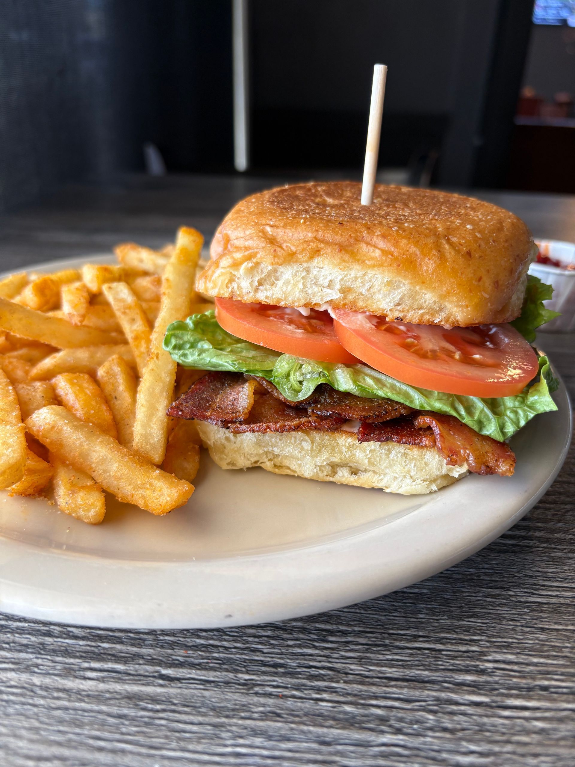 Burger with bacon, lettuce, tomato, and fries on a white plate, next to a small dish of ketchup.
