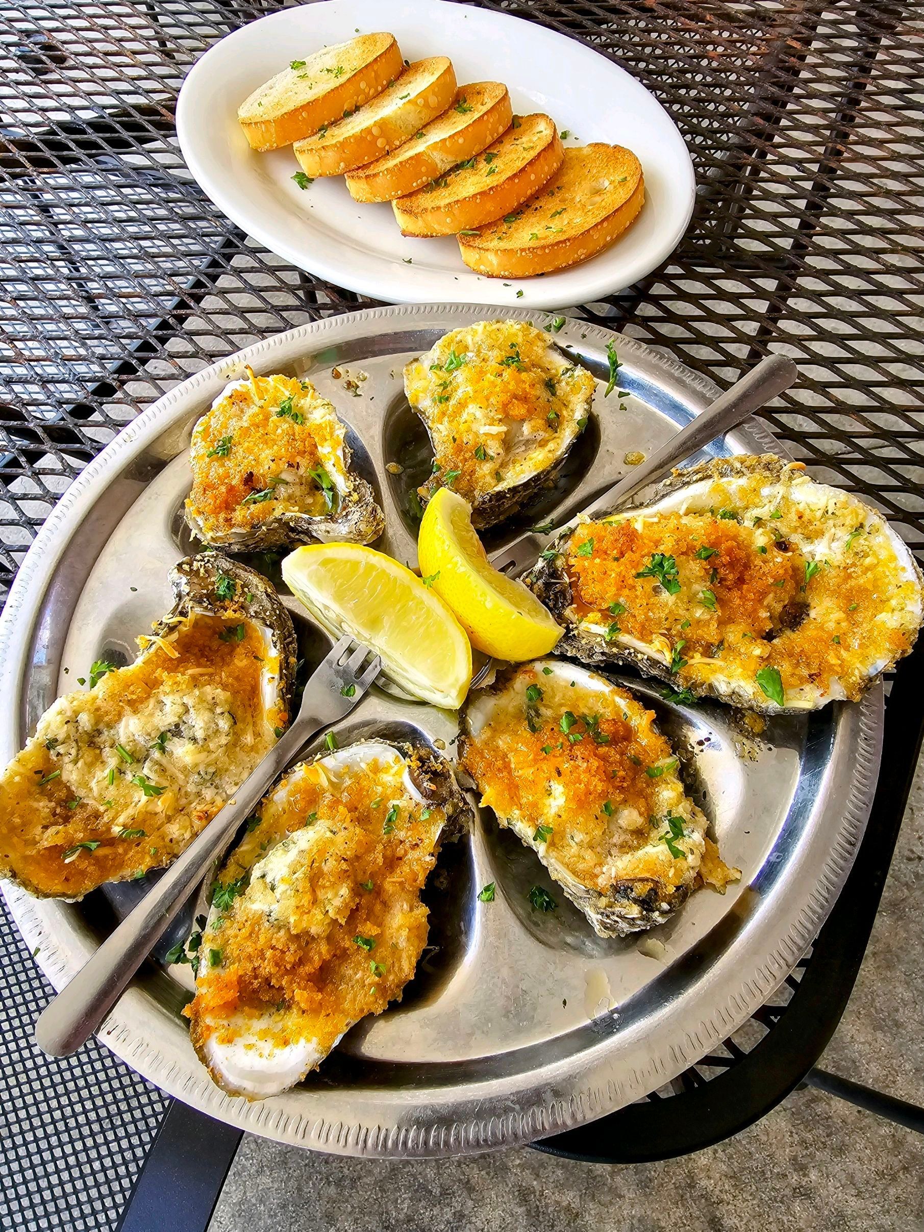 Baked oysters on a metal plate with lemon slices, and garlic bread.