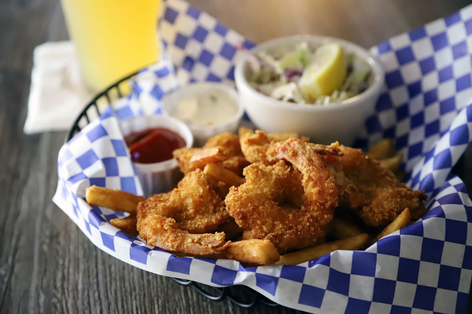 Fried shrimp and fries in a basket with sauces, coleslaw, and drink on a table.