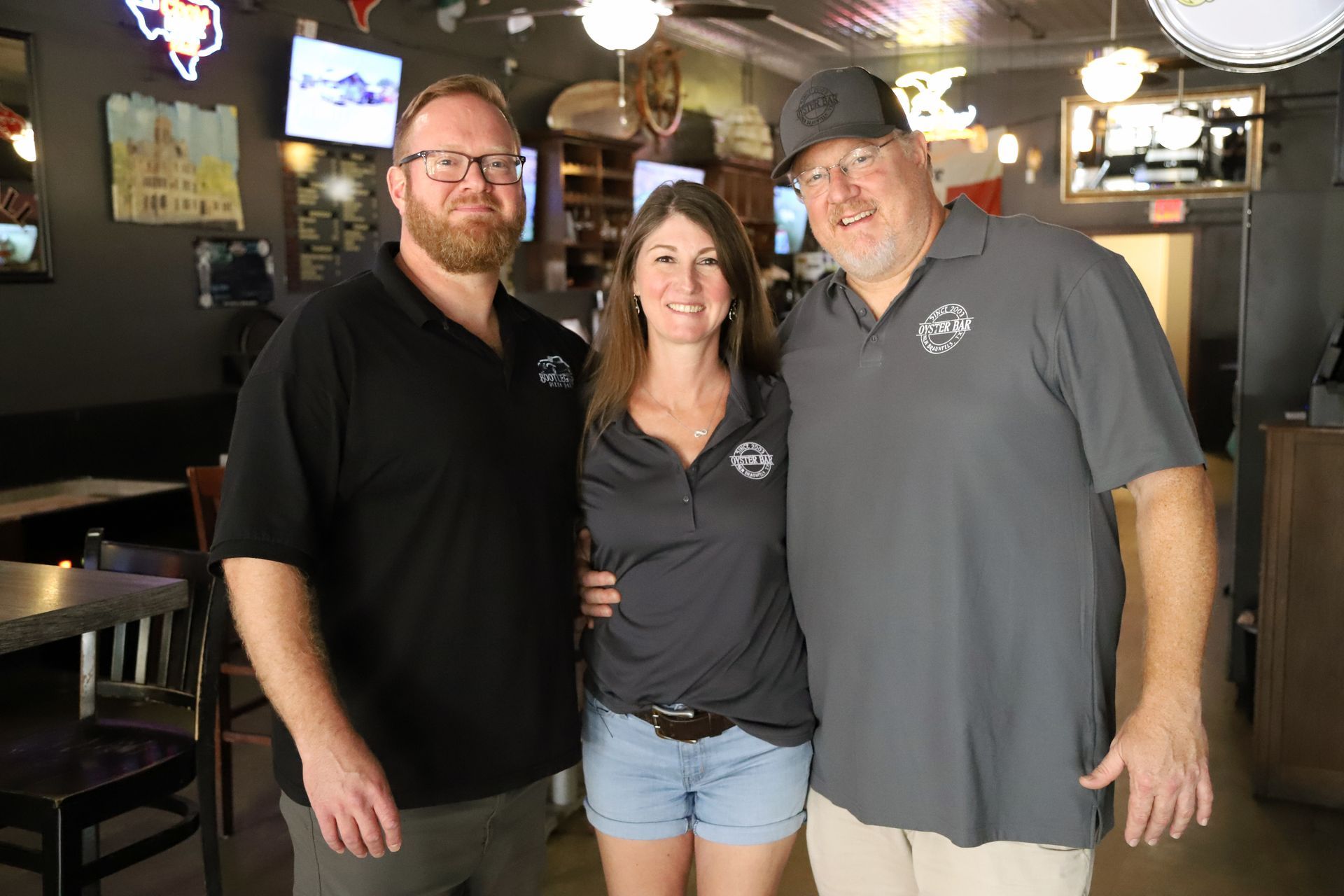 Three people pose at a bar. Two men and a woman smile, wearing dark shirts.