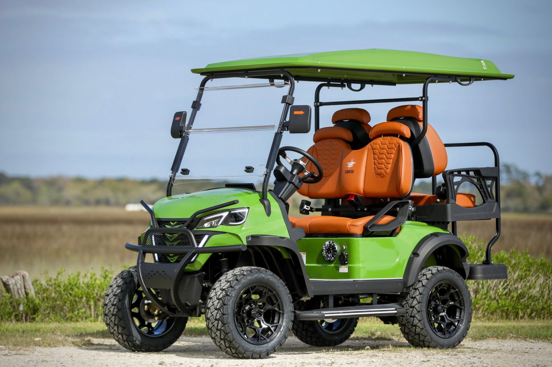 Green off-road utility cart with orange seats on a dirt path in a grassy field, under a clear sky