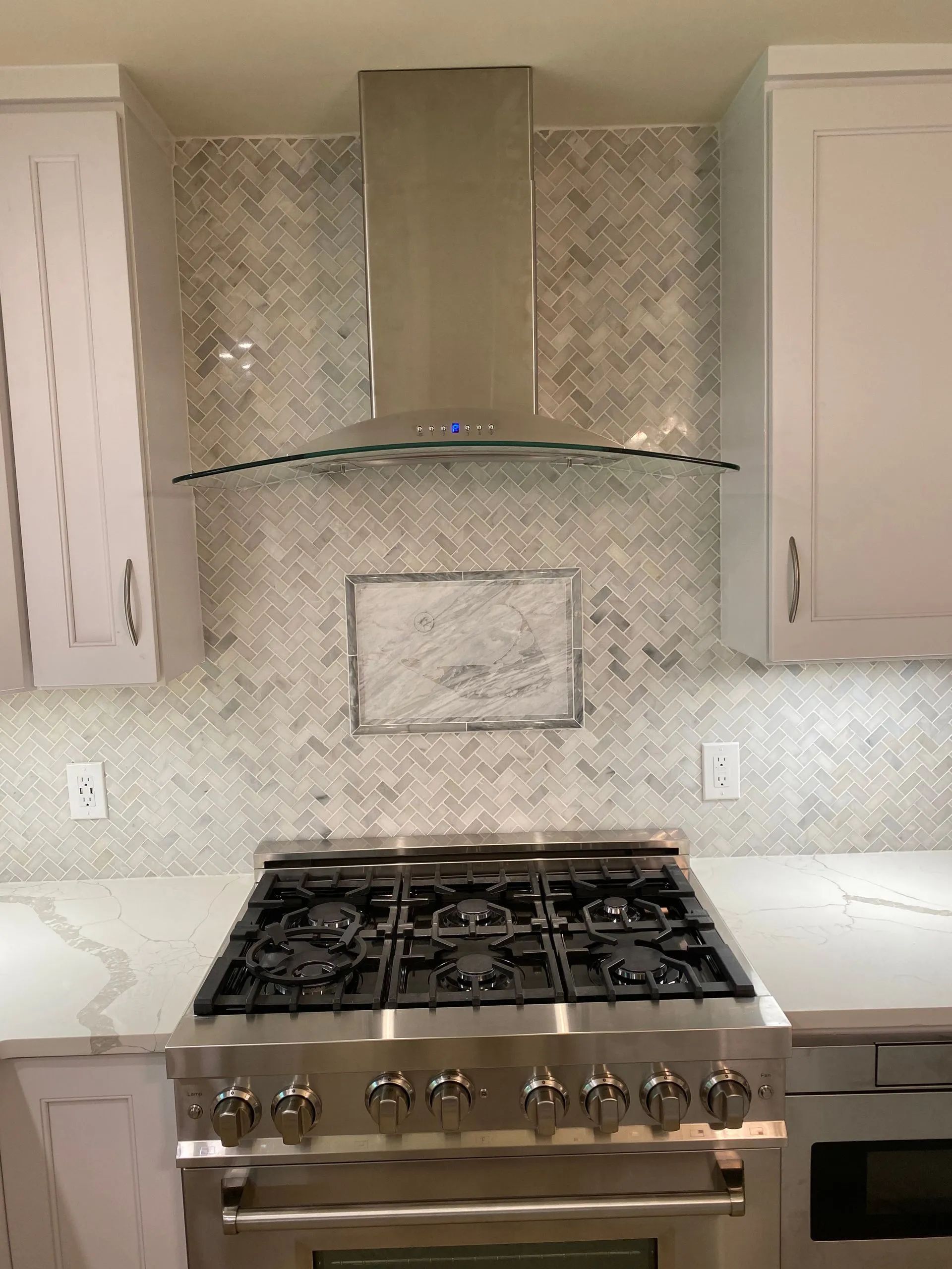 Stainless steel range with a range hood in a white kitchen, herringbone tile backsplash.