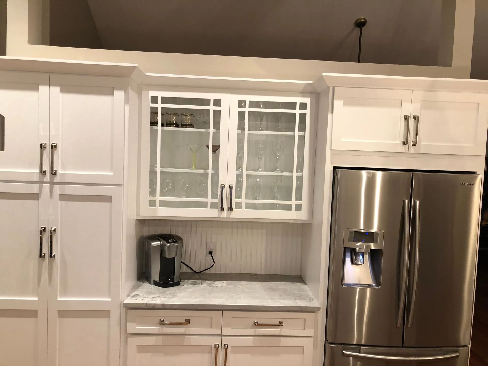 White kitchen cabinets, stainless steel refrigerator. Coffee maker on countertop below glass-front cabinets.