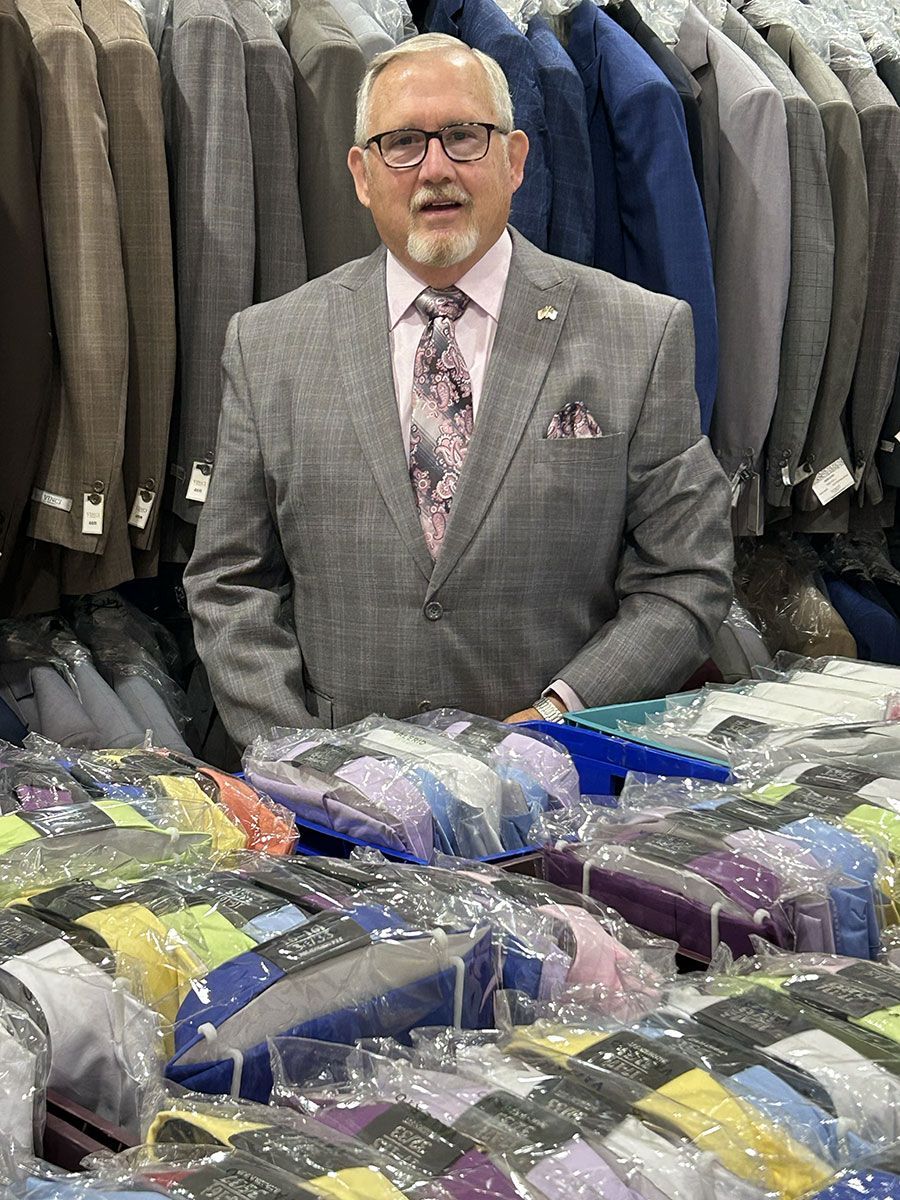 Man in a grey suit stands behind a table of shirts. Suits hang in the background.
