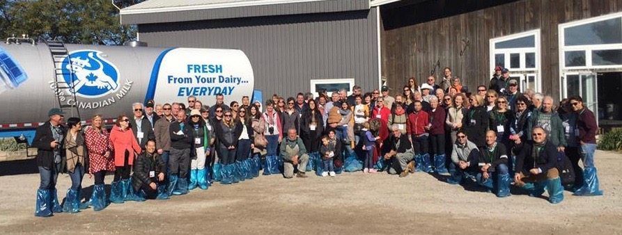 Large group of people pose for a photo in front of a dairy truck and building. Everyone wears blue shoe covers.