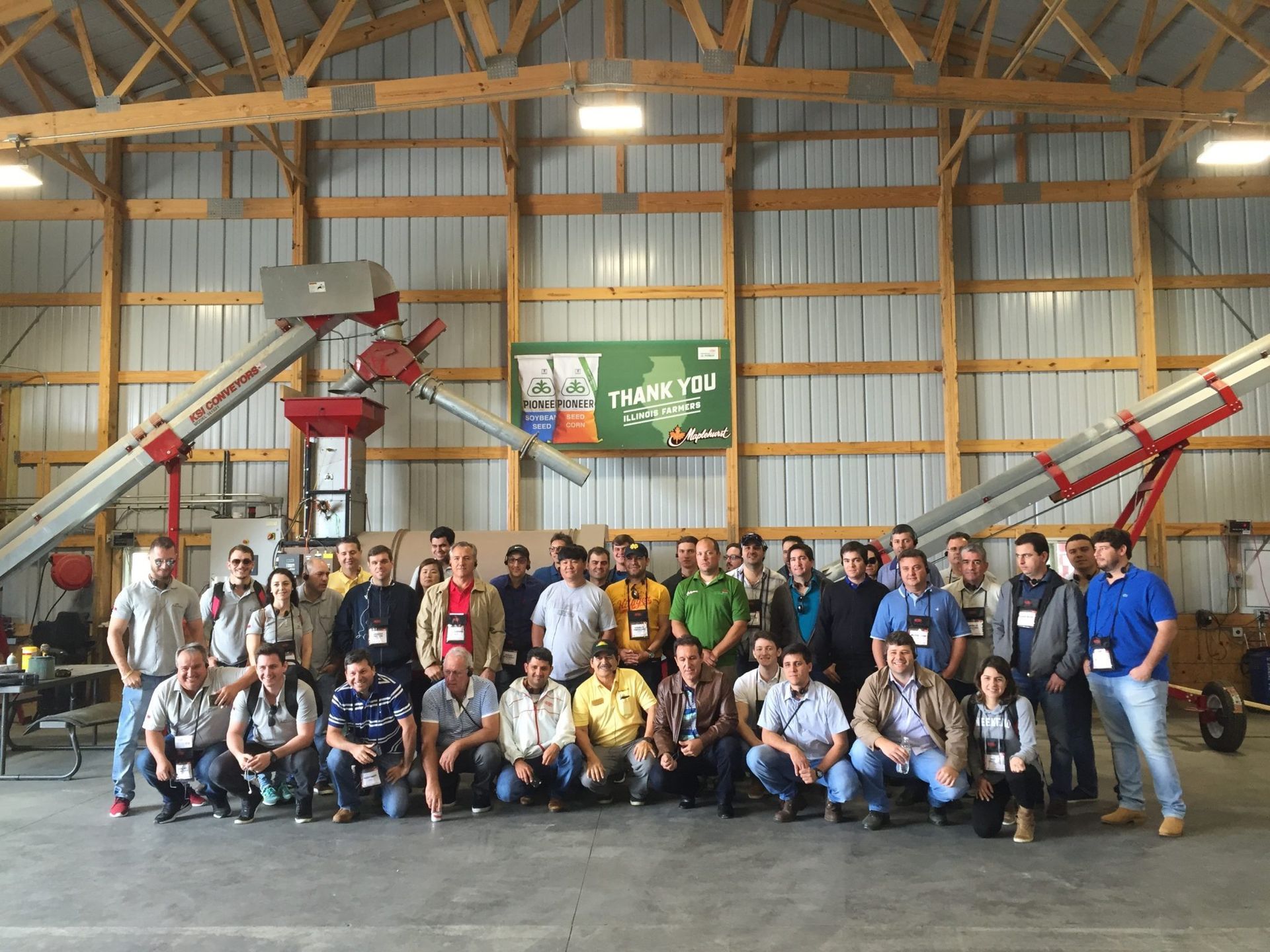 Group of people posing in a barn, standing and kneeling near agricultural equipment, in front of a green sign.
