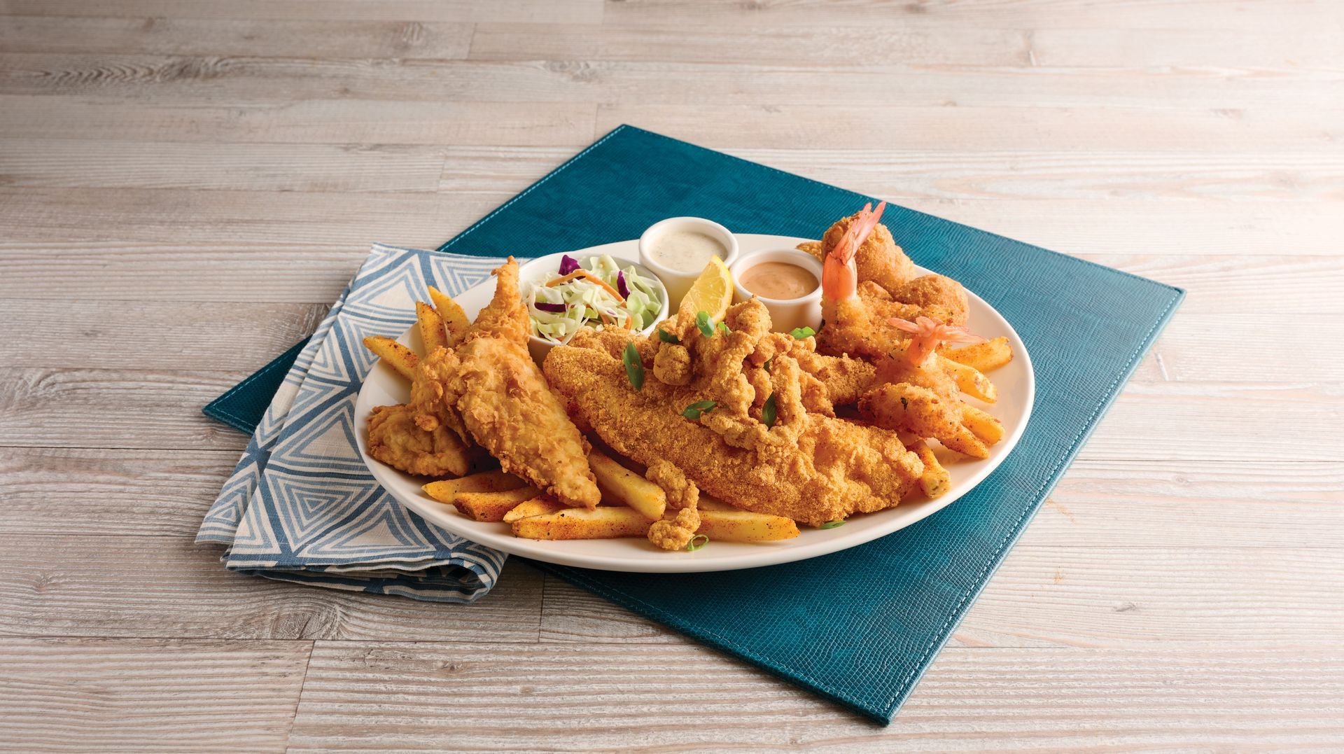 Plate of golden fried fish and shrimp with dipping sauce on a blue placemat on a wooden table