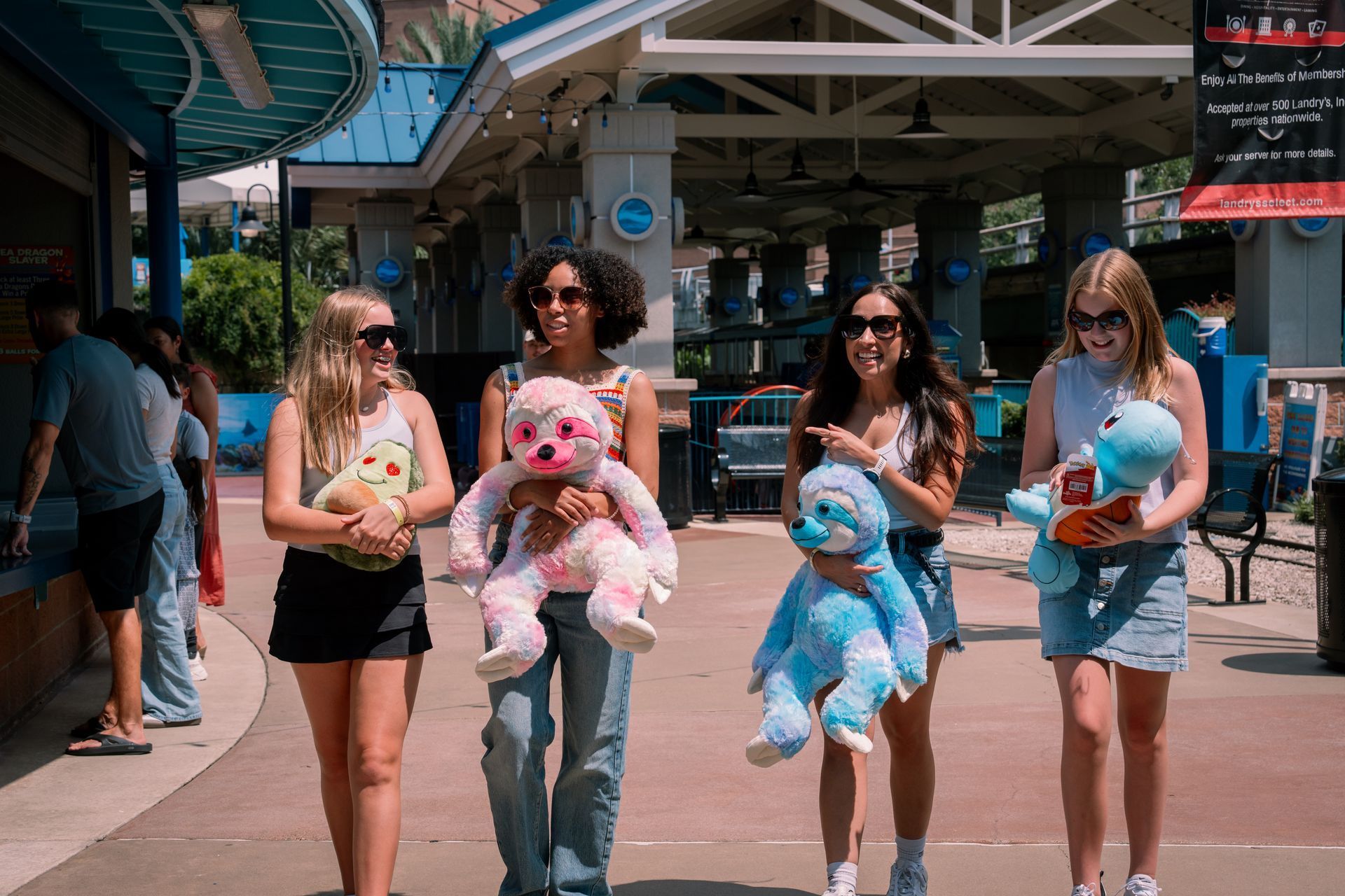 Four people walk, each holding a large plush toy, outside a building with shops.